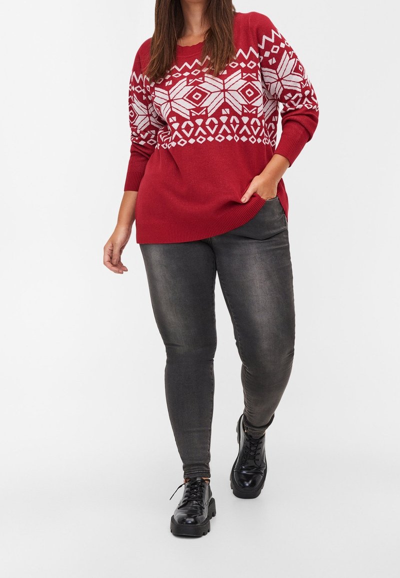 Femme portant un pull rouge à motifs, un jean skinny gris foncé et des chaussures noires à lacets, debout avec une main dans la poche.