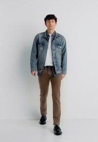 Νέος άνδρας περπατά προς τα εμπρός φορώντας τζιν μπουφάν, λευκό μπλουζάκι, καφέ παντελόνι και μαύρα παπούτσια σε απλό λευκό φόντο.