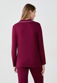 Haut long à manches maroon avec un col en V, doté d'un passepoil blanc le long du col. Tissu lisse et extensible avec une coupe décontractée.