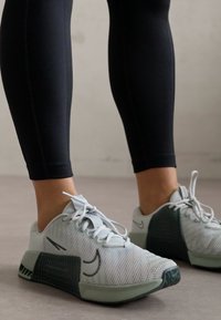 Világosszürke sportcipők, textúrázott hálós felsőrésszel, zöld kiegészítőkkel és tartós talppal; fekete, testre simuló leggingszel párosítva.