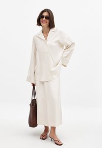 Krämfärgad löst sittande blus med notch-krage och vida ärmar, tillsammans med en matchande lång kjol. Accessoarer inkluderar solglasögon och en vävd väska.