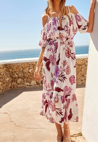 Robe maxi blanche à fleurs avec des motifs de fleurs violettes et rouges, design à épaules dénudées et ourlets volantés. Portée avec des sandales à lanières.
