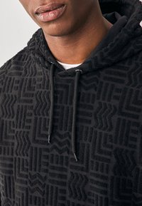 Svart hoodie med ett strukturerat geometriskt mönster. Har en huva med dragsko och ett mjukt, plyschigt material. Grå accenter på mönsterdesignen.