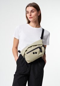 Umhängetasche aus hellbeigem Stoff, mit einer Frontreißverschlusstasche, schwarzen Akzenten und einem minimalistischen Design. Getragen mit schwarzen, strukturierten Hosen.