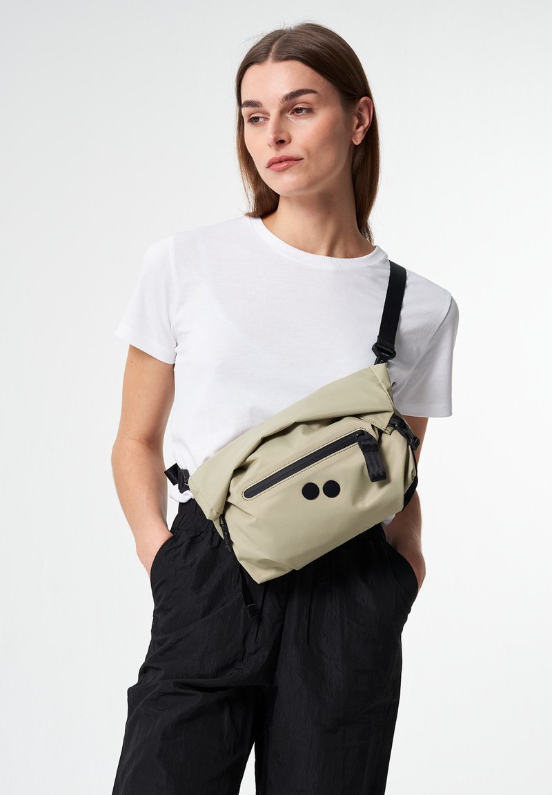 Umhängetasche aus hellbeigem Stoff, mit einer Frontreißverschlusstasche, schwarzen Akzenten und einem minimalistischen Design. Getragen mit schwarzen, strukturierten Hosen.