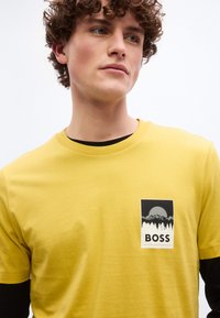 Jeune homme aux cheveux bouclés regardant vers la droite, portant un T-shirt jaune superposé à un haut à manches longues noir, avec un logo "BOSS" représentant une montagne et une forêt.