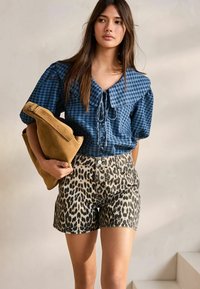 Chemise à carreaux bleue avec un nœud à l'avant, associée à un short beige imprimé léopard. Tient un sac en suede beige.