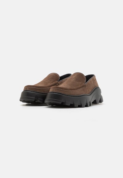 Braune Wildlederschlupfschuhe mit einer dicken schwarzen Gummisohle, die eine abgerundete Zehe und strukturierte Details entlang des Randes aufweist.