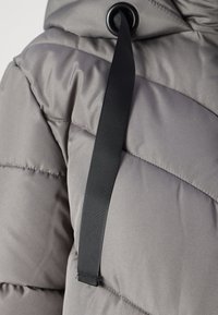 Doudoune grise avec sections matelassées horizontales, une texture lisse et un cordon noir avec un œillet pour les ajustements.