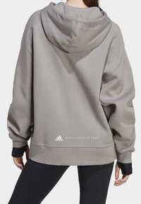 Sudadera gris de gran tamaño con capucha ajustable con cordones, puños acanalados y un logo en el dobladillo. Textura suave y corte holgado, combinada con leggings negros.