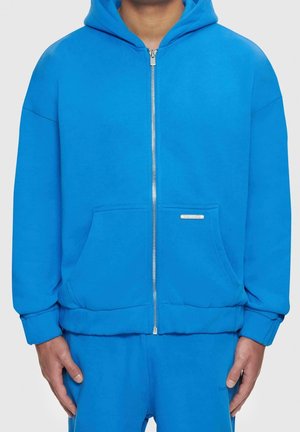 Helle blaue Zip-Hoodie aus weichem Material, mit einer großen Fronttasche und gerippten Bündchen. Schlichter Design mit einem kleinen Markenzeichen auf der Tasche.