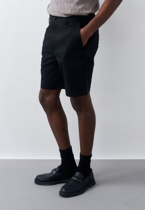 Άνδρας που φορά μαύρο σορτς, μαύρες κάλτσες και μαύρα μοκασίνια, όρθιος με το ένα χέρι στην τσέπη μπροστά σε απλό γκρι και λευκό φόντο.