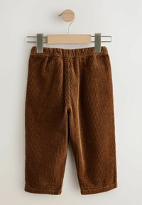 Pantaloni in corduroy marroni con texture a righe verticali, vita elastica e senza tasche, appesi a una clip di legno con una parte superiore rotonda in legno.
