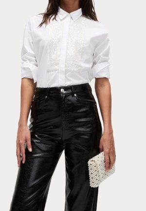 Femme portant une chemise blanche à manches longues avec un panneau avant en sequins, un pantalon en cuir noir taille haute, tenant une pochette ornée de perles dans sa main droite.