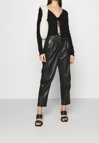 Schwarzes, geripptes Langarmoberteil mit Knoten vorne, kombiniert mit hoch taillierten schwarzen Lederhosen. Geschlossene schwarze Plateau-Sandalen.