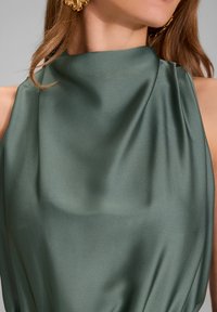 Haut satin vert foncé avec un col montant, présentant un tissu doux et fluide ainsi qu'un léger plissage sur un côté. Des boucles d'oreilles dorées accentuent le look.