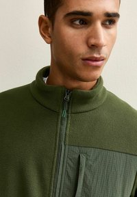 Chaqueta de forro polar verde con cuello alto, frente con media cremallera y panel texturizado en el pecho. Incluye bolsillo con cremallera y detalles de tela suave.