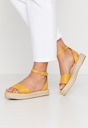 Pieds portant des sandales espadrilles jaunes à brides chevilles avec semelles compensées en jute tressé et pantalon blanc raccourci sur un fond uni.