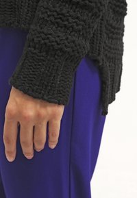Maglione nero lavorato a maglia con un motivo testurizzato, mostrato accanto a un tessuto blu. La manica presenta un polsino a coste e un design morbido.