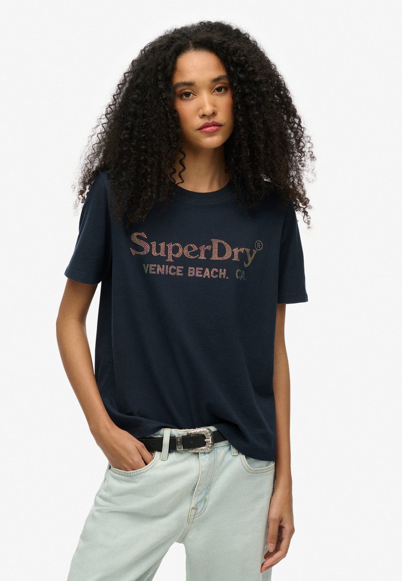 Mørkeblå bomulds t-shirt med "SuperDry" og "Venice Beach" i farverig tekst, korte ærmer, afslappet pasform, kombineret med lyse denim jeans.