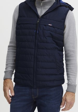 Homme portant une veste matelassée sans manches bleu marine par-dessus un pull gris zippé et un jean, debout les bras détendus le long du corps.