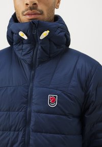 Námořnická pufovaná bunda s vysokým límcem, zipem a dvěma bílými knoflíky. Má vyšívané logo na hrudi a prošívanou texturu.