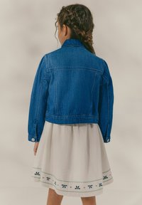 Chaqueta de mezclilla con corte clásico en azul oscuro, con cuello de punta y puños abotonados, combinada con una falda blanca adornada con bordados verdes.
