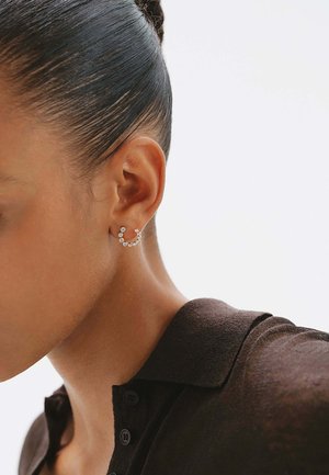 Mujer con cabello oscuro recogido en un moño, que lleva una camisa marrón con cuello y un pequeño aro con piedras transparentes en la oreja izquierda.