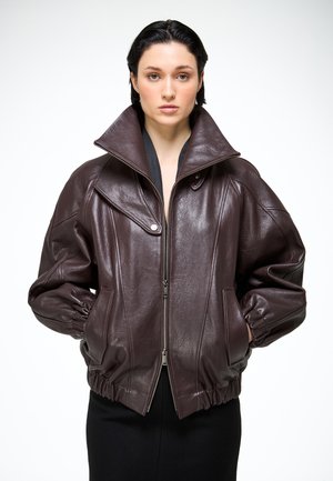 Mladá žena v nadrozmernej tmavohnedej koženej bunde s vysokým golierom, zipsom a rukami v vreckách stojaca na bielom pozadí.