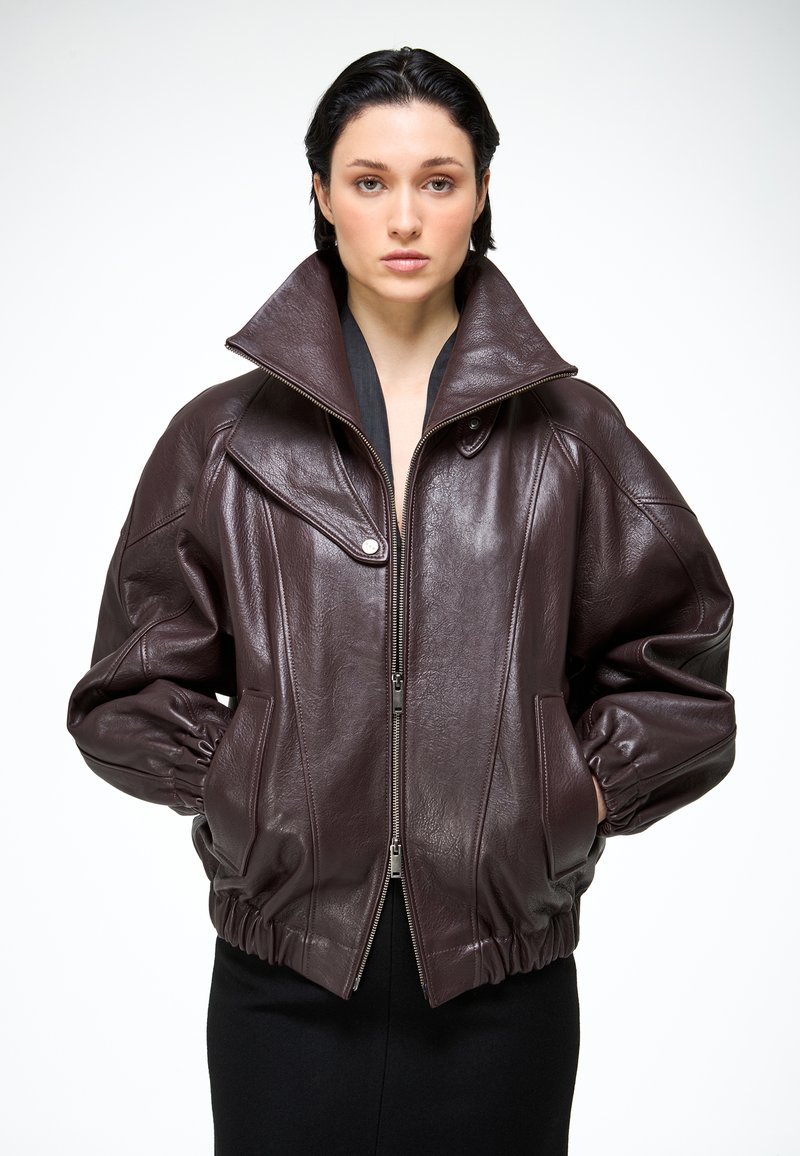 Mladá žena v nadrozmernej tmavohnedej koženej bunde s vysokým golierom, zipsom a rukami v vreckách stojaca na bielom pozadí.