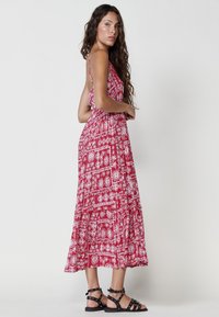 Rotes Maxikleid mit weißen geometrischen Mustern, Spaghetti-Trägern, tailliertem Oberteil und gestuftem Rock. Getragen mit schwarzen, genagelten Sandalen.