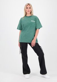 Groen oversized t-shirt met een vervaagde textuur en logo op de borst, gecombineerd met zwarte cargobroek met zijzakken en gestrikte accenten.
