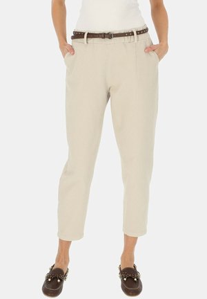 Kobieta ubrana w beżowe, krótkie spodnie z kieszeniami, brązowy pas z ćwiekami oraz ciemnobrązowe mokasyny z ozdobnymi dodatkami.