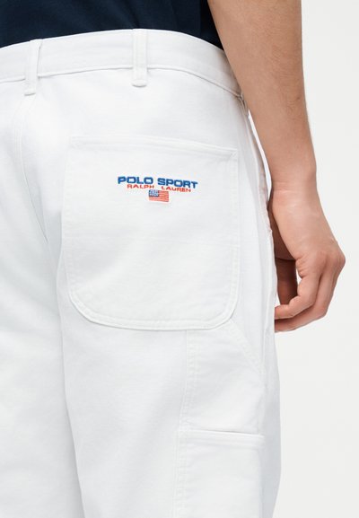 Κοντινό πλάνο στην τσέπη λευκών σορτς με κέντημα λογότυπου "Polo Sport Ralph Lauren" και αμερικανικής σημαίας πάνω στο ισχίο ενός ατόμου, με εμφανή το χέρι.