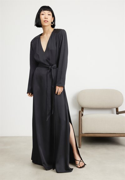 Μαύρο φόρεμα με ζώνη που έχει μακριά μανίκια, χαμηλό ντεκολτέ, ζώνη στη μέση και πλαϊνό σκίσιμο. Λείο ύφασμα με ρέουσα σιλουέτα. Μαύρες σαγιονάρες με ιμάντες.