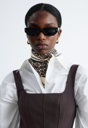 Sonnenbrille mit einem schwarzen rechteckigen Rahmen, kombiniert mit einem geometrisch gemusterten Schal in Beige- und Dunkeltönen, getragen über einem weißen Hemd und einem braunen Kleid.