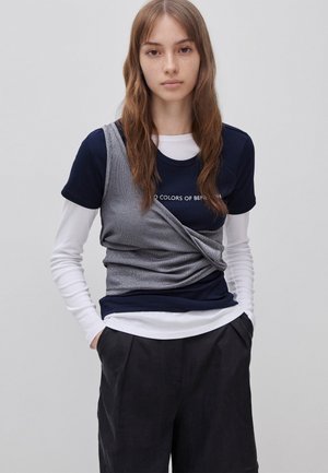 Jonge vrouw met lang bruin haar, die gelaagde marineblauwe en witte lange mouwen draagt met een grijs wikkeldetail en een zwarte broek, staand met handen in de zakken.