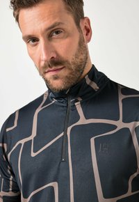 Mann mit kurzem Bart, der ein schwarz-beiges, gemustertes Sportoberteil mit halbem Reißverschluss trägt und mit neutralem Gesichtsausdruck direkt in die Kamera schaut.