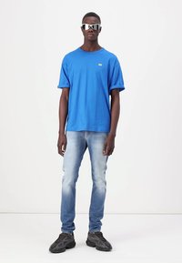 Camiseta de algodón azul con mangas cortas y un pequeño logotipo verde, combinada con jeans azul claro y zapatillas negras robustas. Gafas de sol reflectantes.