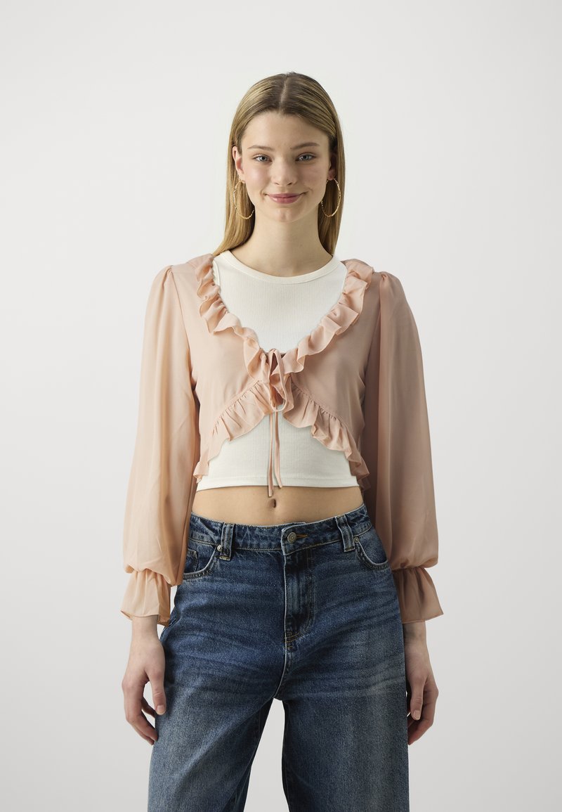 Light pink, sheer, long-sleeve blouse with ruffled front and tie closure, layered over a white fitted top, paired with high-waisted blue jeans.