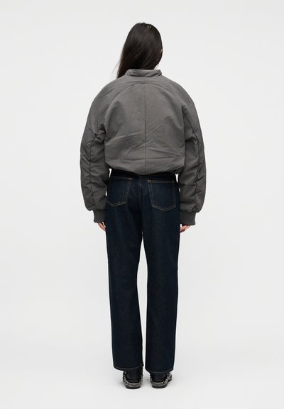 Άτομο με μακριά σκούρα μαλλιά που φοράει γκρι μπουφάν με πούπουλα, σκούρα μπλε τζιν που είναι ίσια στο πόδι και μαύρα παπούτσια με αλυσίδες, στέκεται γυρίζοντας την πλάτη.