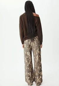 Maglione off-shoulder marrone scuro abbinato a pantaloni a gamba larga in camouflage con motivi beige e verde olivo. Tessuto liscio e testurizzato.