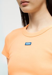 Γυναίκα που φοράει ροδακινί κοντομάνικο πουκάμισο με ένα μικρό μπλε λογότυπο "HUGO" κοντά στην κλείδα.