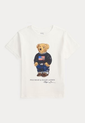 Hvit bomull T-skjorte med et grafisk print av en teddybjørn iført en marineblå genser med amerikansk flagg, blå jeans og brune sko.