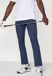 Μπλε σκούρο παντελόνι γκολφ κατασκευασμένο από ελαφρύ υλικό, με ραμμένη εφαρμογή και απαλή υφή. Συνδυάζεται με λευκά αθλητικά παπούτσια και γάντι γκολφ.