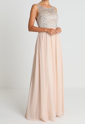 Kvinde iført ærmeløs gulvlængde kjole med palietter på overdelen og flydende beige nederdel, stående mod en ensfarvet lys baggrund.