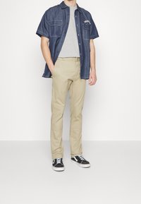 Man wearing beige pants, grey t-shirt, unbuttoned dark blue short-sleeve denim shirt, and black-and-white sneakers against white background.