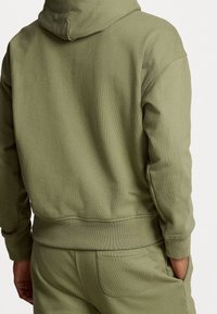Olivově zelená mikina s kapucí vyrobená z měkkého materiálu, s ribbaným lemem a manžetami, volným střihem a šitými švy.