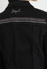 Musta takki takana, jossa on brodeerattu "Desigual"-logo ja vaaka koristekaistale, jossa on punaisia ja valkoisia geometrisia kuvioita.