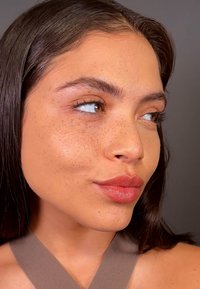 Nærbilde av et ansikt med mellomfarget hud, synlige fregner, langt mørkt hår, fyldige lepper med rosa glans, og iført en brun ermeløs topp.